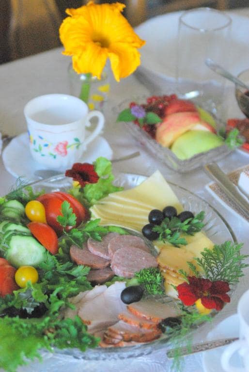 A beautifully arranged platter features an assortment of fresh vegetables, cured meats, and a selection of cheese. Colorful tomatoes, cucumbers, and olives are complemented by leafy greens and herbs. A delicate flower adds a touch of brightness to the white table setting.