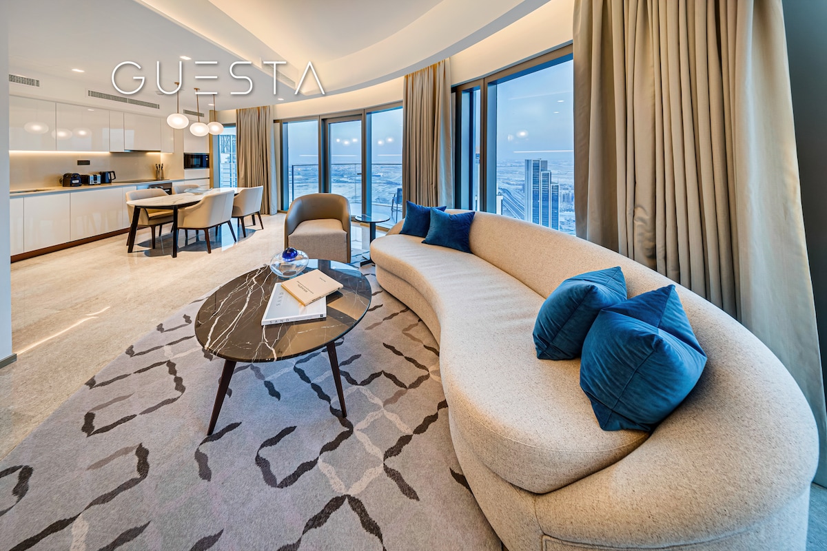 A modern living area is highlighted by a curved sofa adorned with blue cushions, complemented by a sleek coffee table. A dining table with chairs is situated in the background, while large windows frame views of the city skyline.