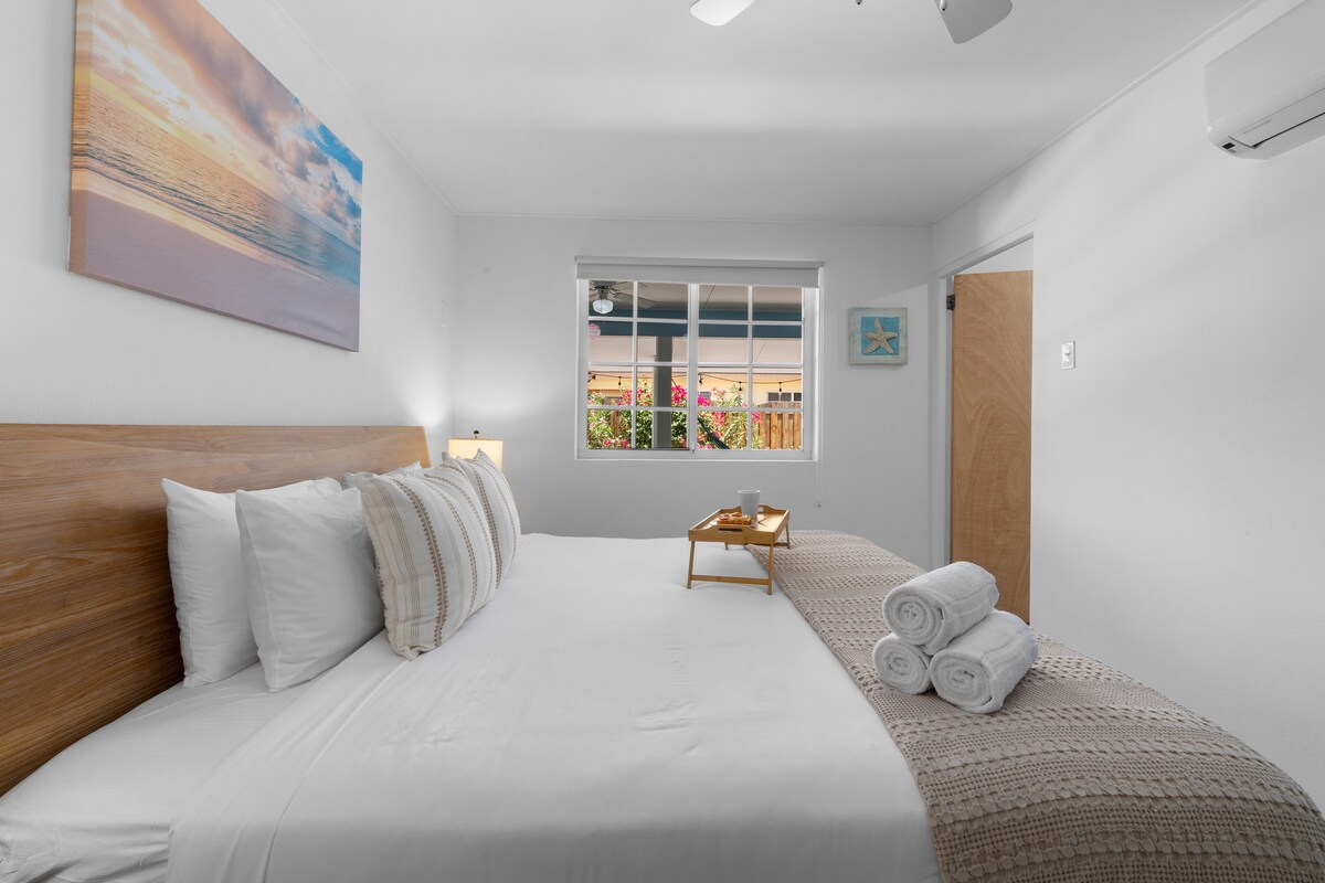 Საძინებელი დიდი საწოლით, თეთრი თეთრეულით, ხის გარნიტურით, საწოლისპირა ლამპებითა და პატიოს ფანჯრის ხედით.