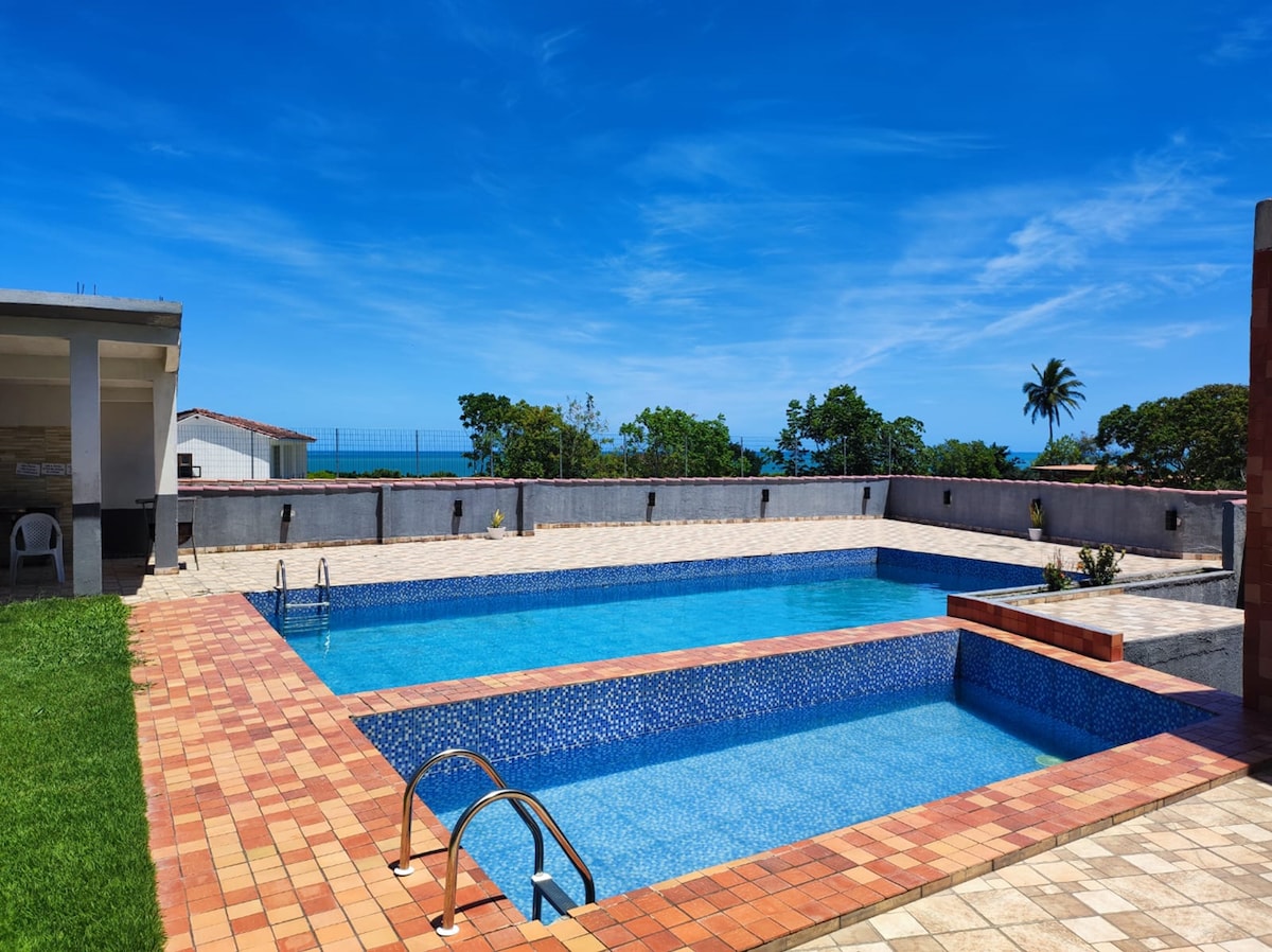 Two inviting swimming pools are set in a sunlit area surrounded by tile. A large open space features manicured grass and tropical trees, with the ocean visible in the background under a clear blue sky.