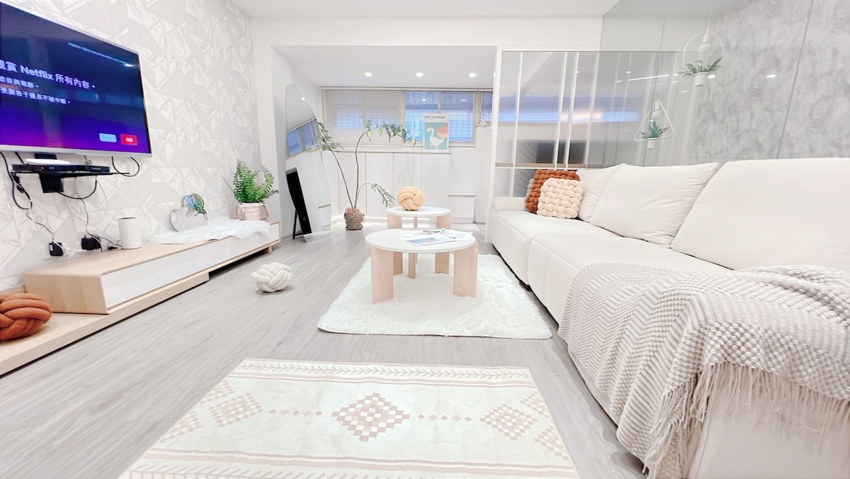 A modern living space features a spacious sectional sofa paired with decorative cushions. A low coffee table is centered on a soft area rug. A large television is mounted on the wall, with natural light pouring in through windows, adding a bright ambiance to the room.