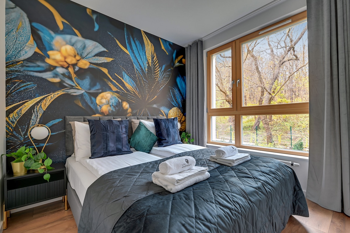 A well-appointed bedroom features two spacious beds dressed in crisp white linens, accented by a quilted dark coverlet. Decorative pillows in shades of teal and emerald are arranged on the beds. A large window offers a view of greenery outside, framed by light gray curtains.