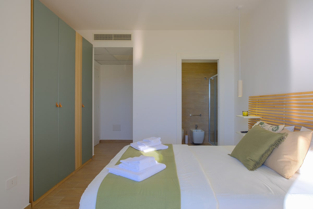 A calming bedroom showcases a spacious bed with light bedding and decorative pillows. A wardrobe with green accents stands on one side, while a modern bathroom is visible through an open door. Warm light fills the room, enhancing its peaceful environment.