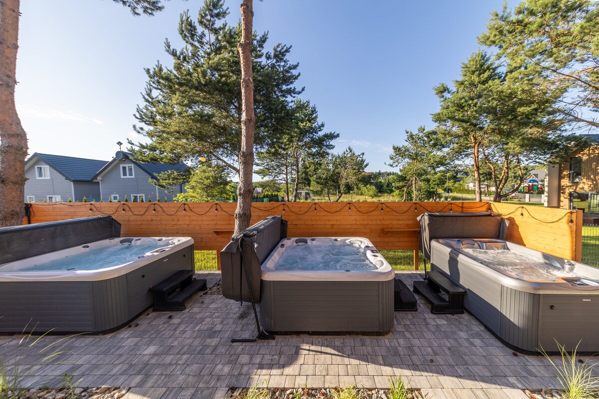 Three outdoor jacuzzis are positioned on a wooden deck, surrounded by lush trees. The hot tubs are separated by wooden fencing and face a serene landscape. Soft lighting enhances the ambiance, making it an inviting space for relaxation.