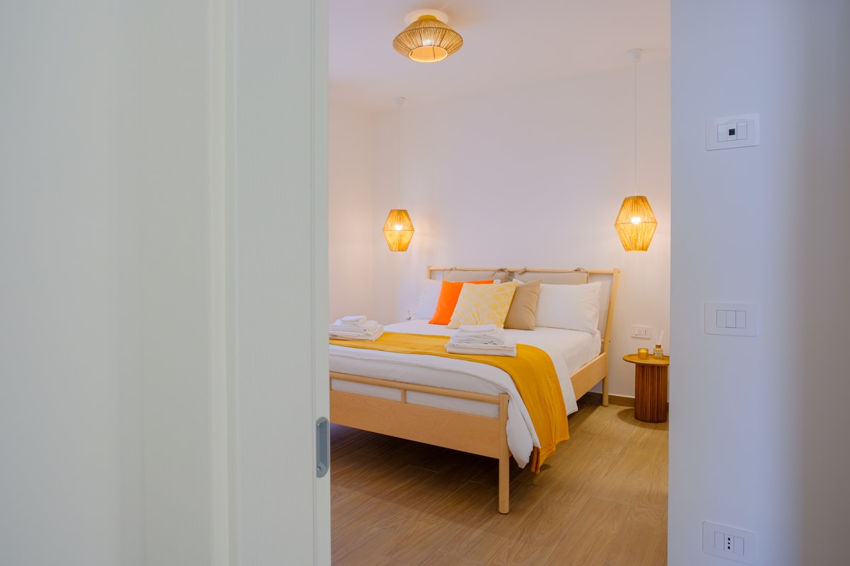 A well-furnished bedroom is viewed from a doorway, featuring a spacious bed with bright orange and white accents. Two pendant lights add a warm glow, while fresh towels are neatly placed on the bed. Natural light fills the room, creating a calming atmosphere.