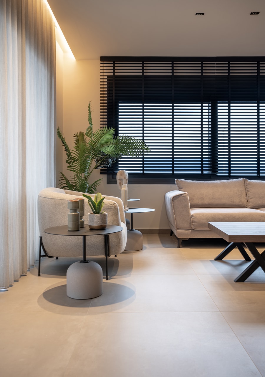 The living area features a comfortable sofa and two armchairs positioned beside small side tables. A potted plant adds a touch of greenery, while light-filtering shades provide privacy. Natural light enters through the window, enhancing the modern decor with clean lines.