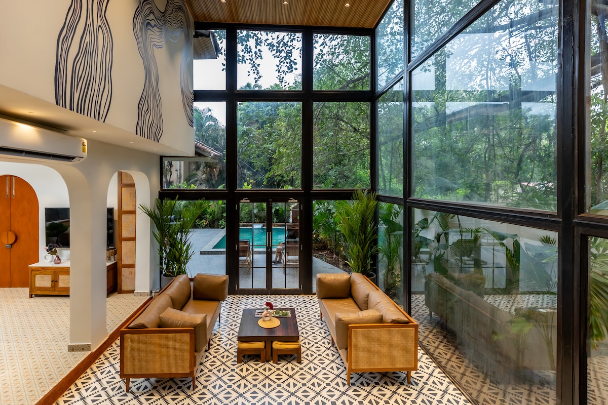 A spacious living area is showcased with large glass walls that offer views of the outdoor pool and greenery. Two comfortable sofas are arranged around a central coffee table, with decorative elements enhancing the contemporary design. Natural light fills the space, creating an airy ambiance.