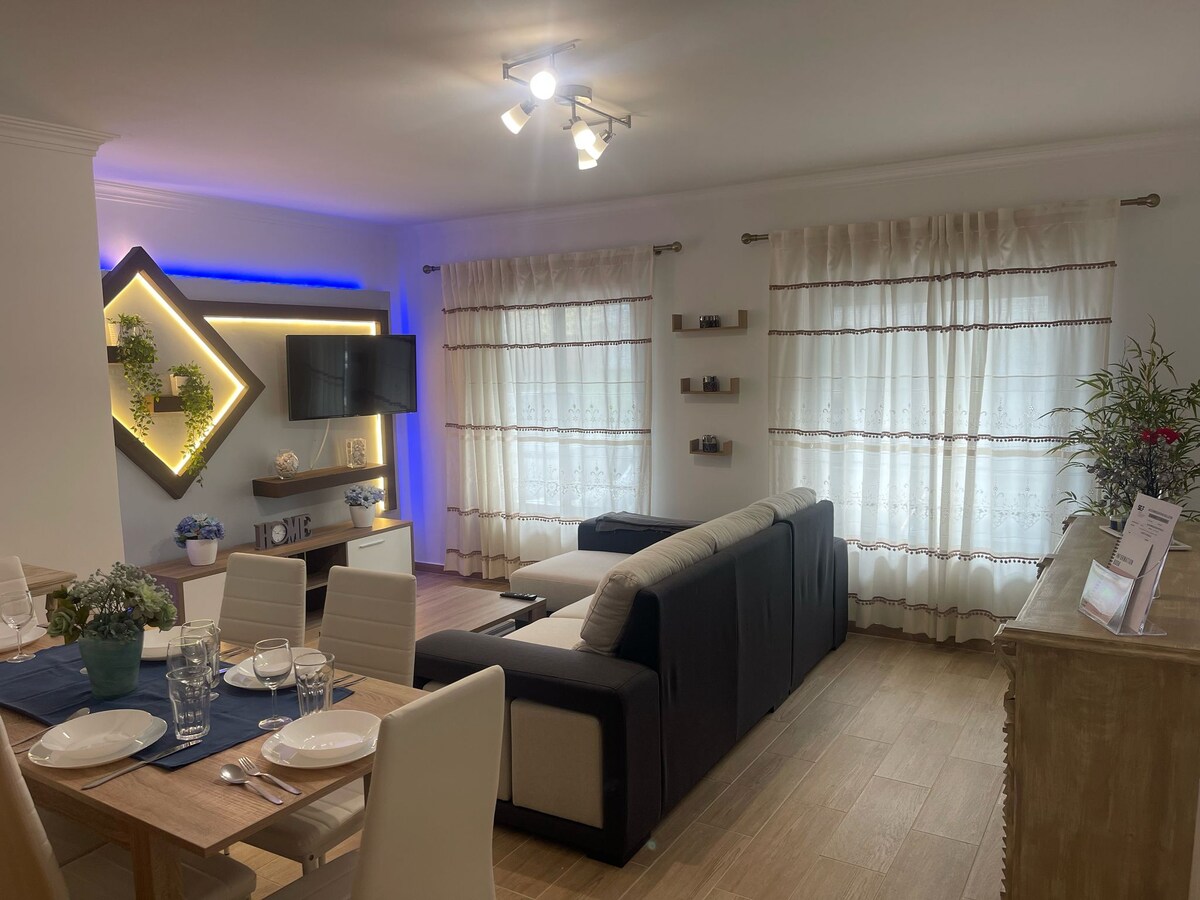 The living area features a modern design with a sectional sofa and a TV mounted on a decorative wall. Large windows with light curtains allow natural light to fill the space. A dining table is set with tableware, accompanied by plants and shelves on the walls.