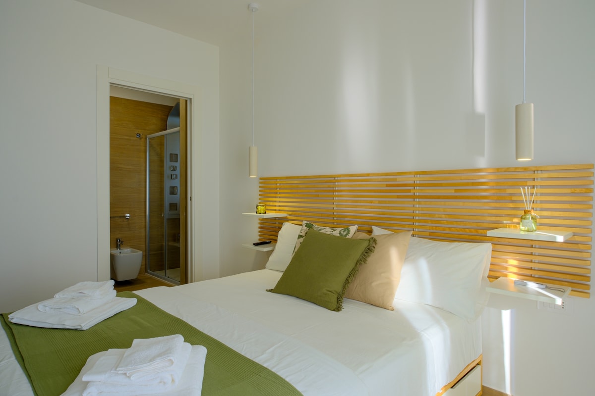 A well-appointed bedroom features a spacious bed with crisp white linens and decorative pillows. Soft lighting is provided by pendant lamps on either side. A modern en-suite bathroom is visible through a doorway, highlighted by warm wood tones and inviting design.