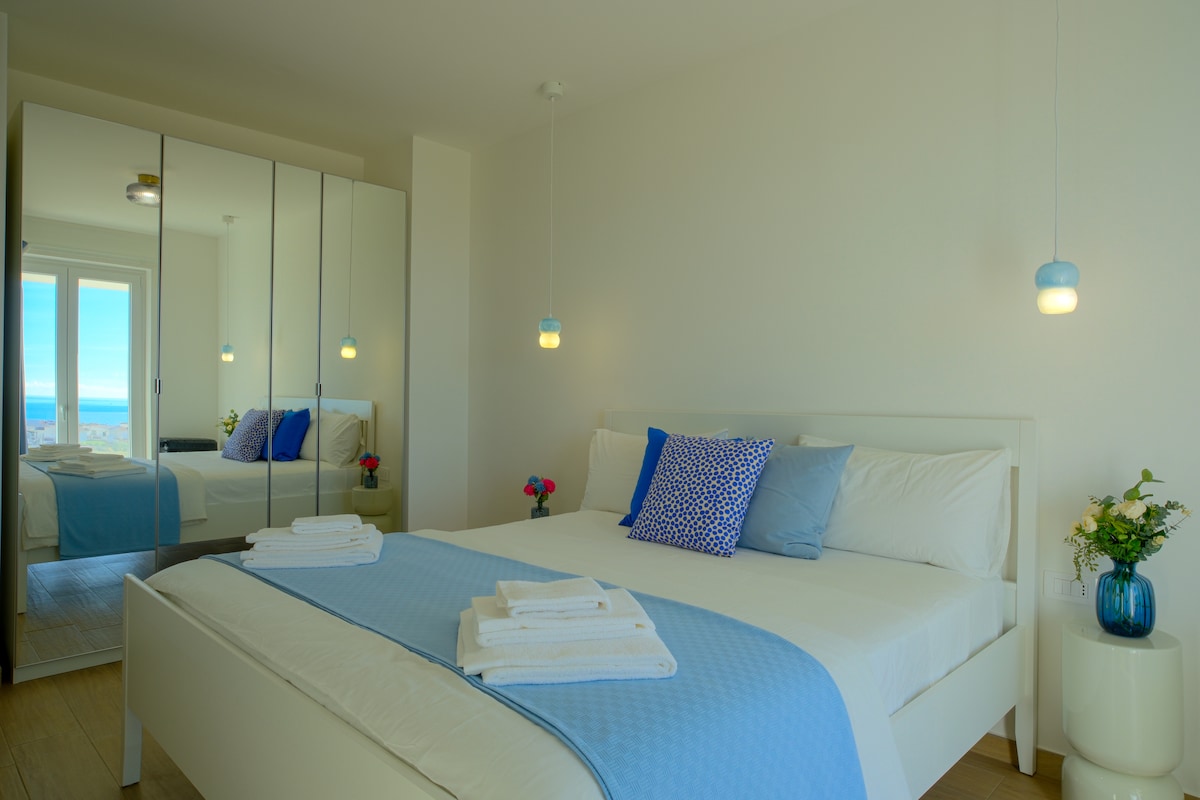 A well-appointed bedroom features a spacious bed with a light blue throw and numerous folded towels. Reflected in a large mirrored wardrobe, soft lighting illuminates the room. A small vase with flowers adds a touch of freshness, while a glimpse of the sea is visible through large windows.