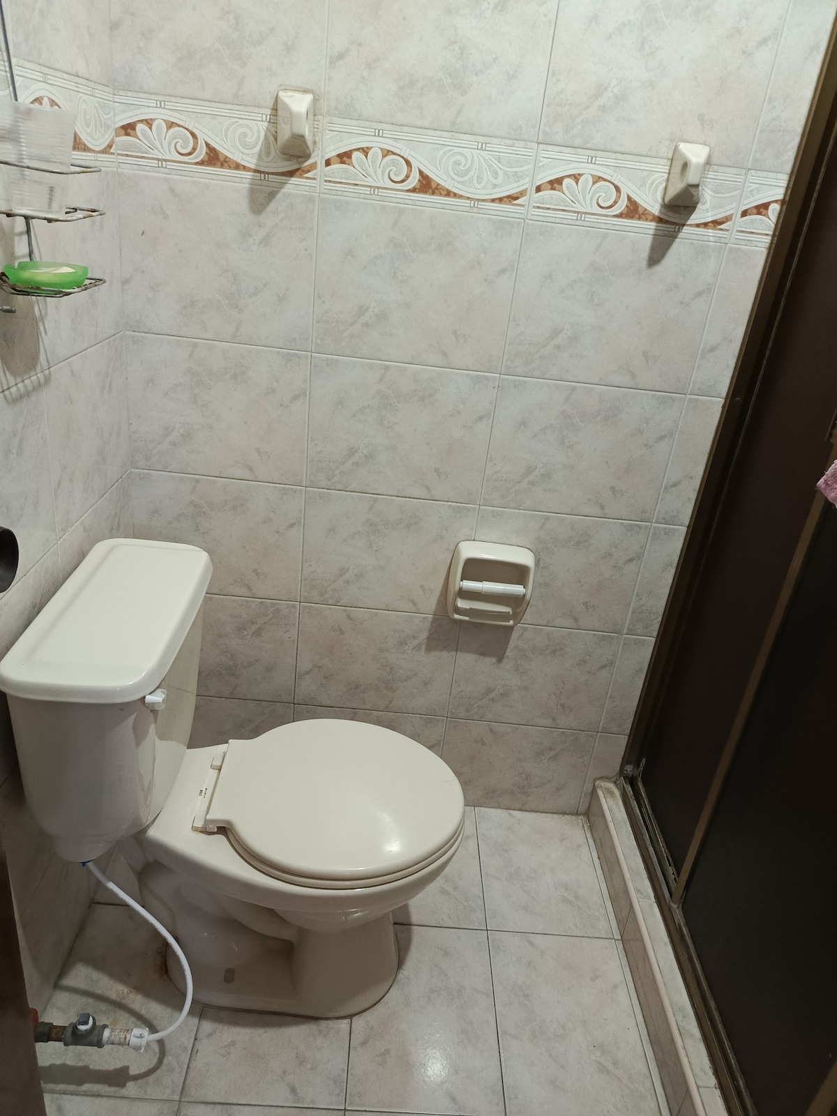 The bathroom is fitted with neutral tiles and features a compact toilet. A shower area is visible on one side, with a sliding door for access. Shelving is installed above the toilet, providing space for toiletries.