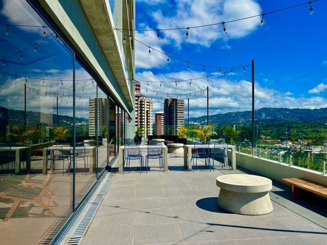 A rooftop terrace features modern furnishings, including a circular concrete table and blue wire chairs. String lights are suspended overhead, while views of the surrounding skyline and hills create a scenic backdrop. Floor-to-ceiling windows provide a panoramic perspective of the outdoor space.
