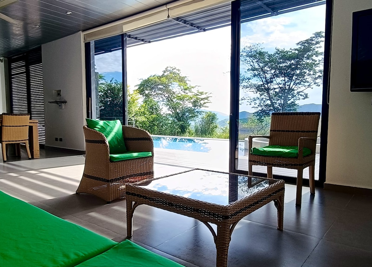 Seating arrangements consist of two green-accented chairs and a glass-topped coffee table, offering a view of the outdoor pool area and lush surroundings through expansive glass doors. Natural light enhances the open, airy feel of the space.