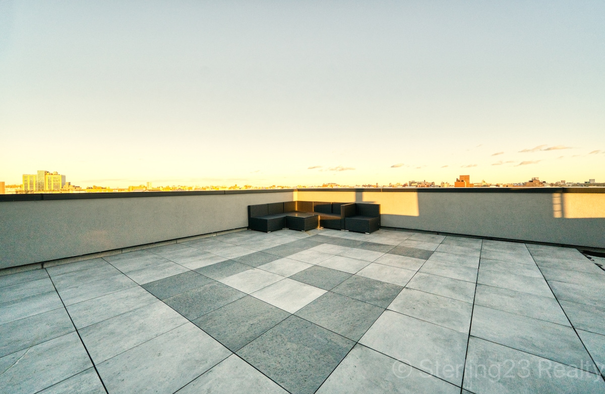 A shared rooftop space showcases a spacious patio area featuring modern seating arrangements. The flooring consists of large square tiles in varying shades. The city skyline is visible in the distance, providing a serene backdrop for relaxing moments.