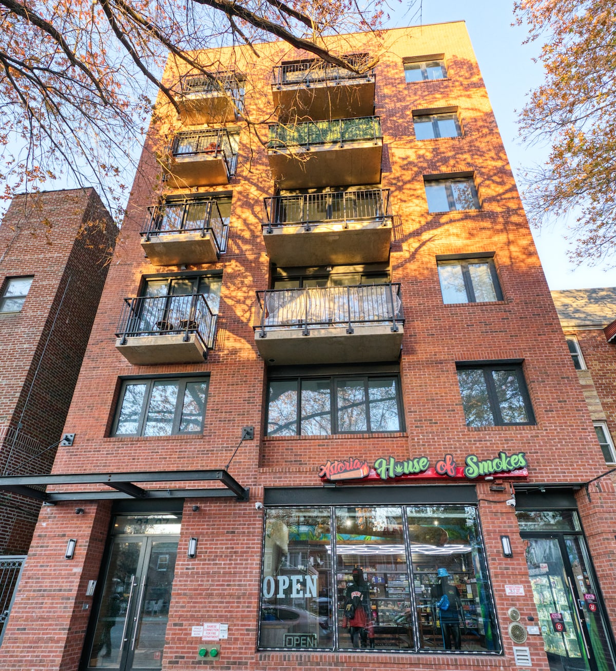 The modern brick building stands five stories high, featuring multiple balconies with black railings. Ground-level storefronts are visible, alongside glass doors adorned with an 'OPEN' sign. Tree branches above add an element of nature to the urban setting.