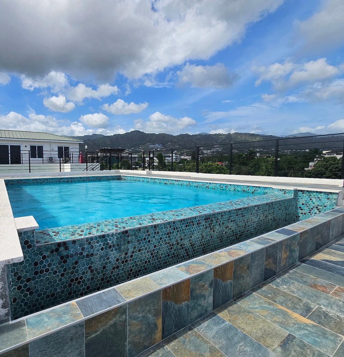 A rooftop pool is showcased, featuring turquoise water and a stone-tiled border. The serene view includes rolling hills and scattered clouds. The pool area is enclosed with a protective railing, blending leisure with safety.