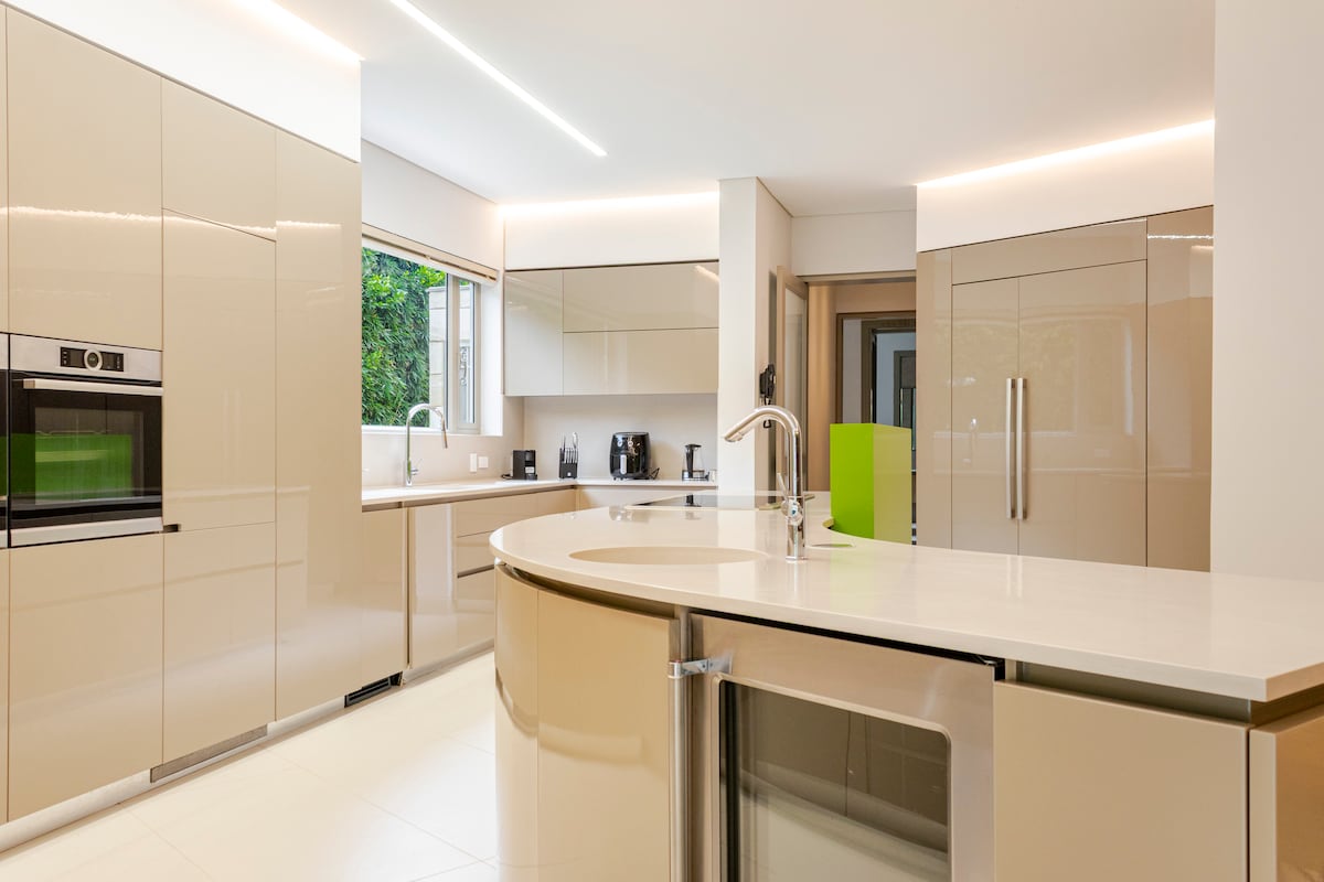 A modern kitchen features sleek, beige cabinets and a spacious island with a built-in sink. Large windows allow natural light to illuminate the room, while kitchen appliances are arranged for convenience. The minimalist design promotes an inviting cooking environment.
