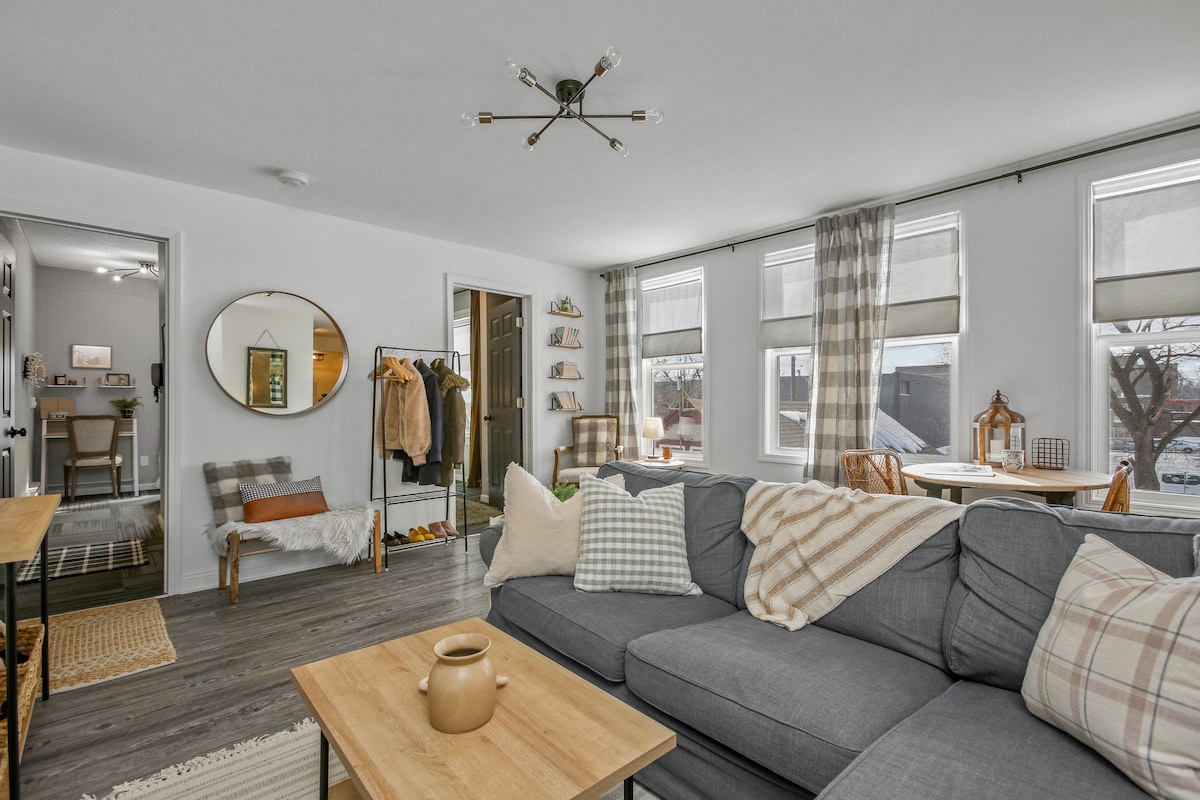 A modern living space showcases a comfortable gray couch adorned with patterned cushions. Light streams through multiple windows dressed in soft curtains. A wooden coffee table sits in front, while a small dining area can be seen in the background, featuring a round table and chairs.