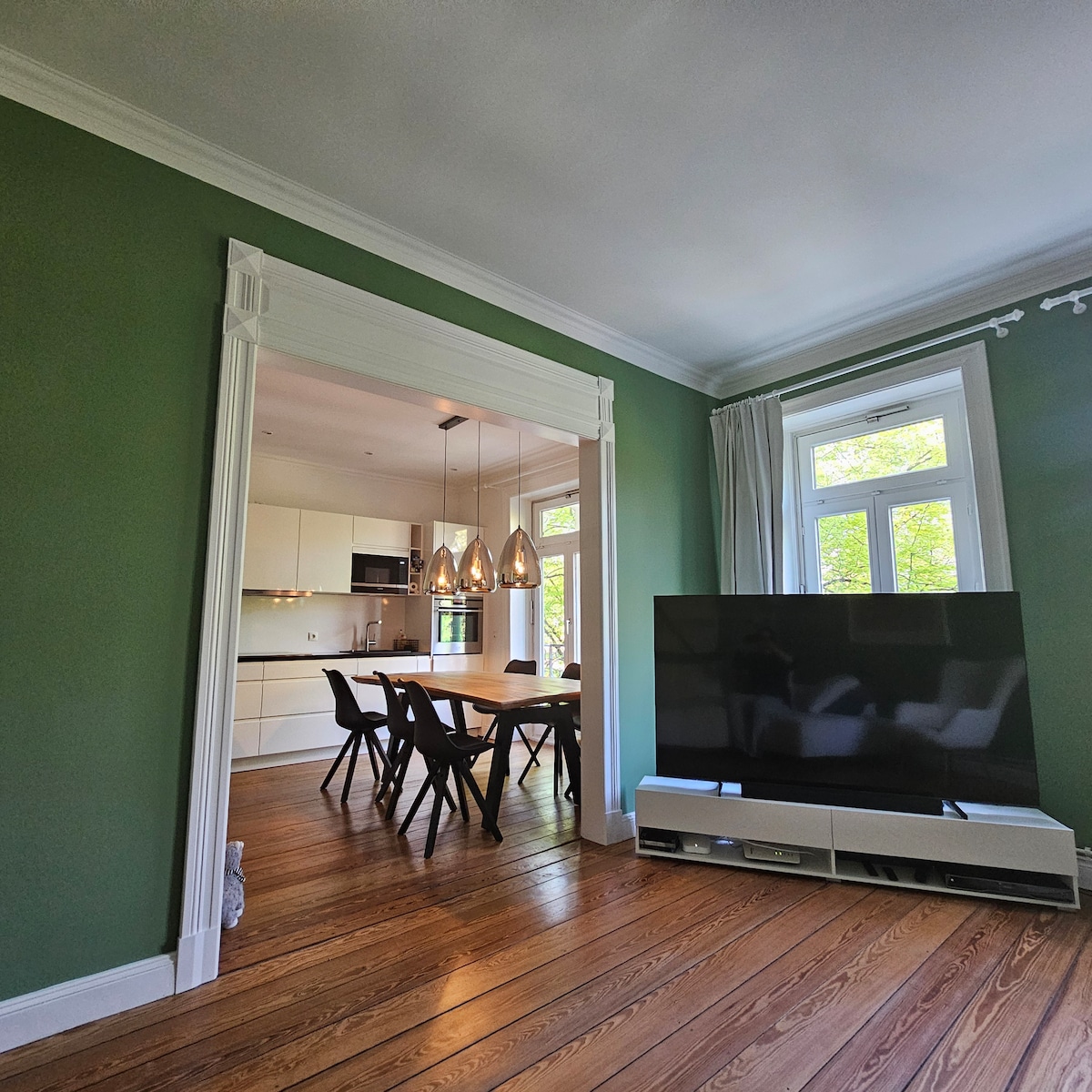 A bright living space is separated from the modern kitchen by an arched doorway. The kitchen features a dining area with a large table and stylish chairs, illuminated by pendant lighting. A large television is positioned on a media console, with natural light filtering through nearby windows.