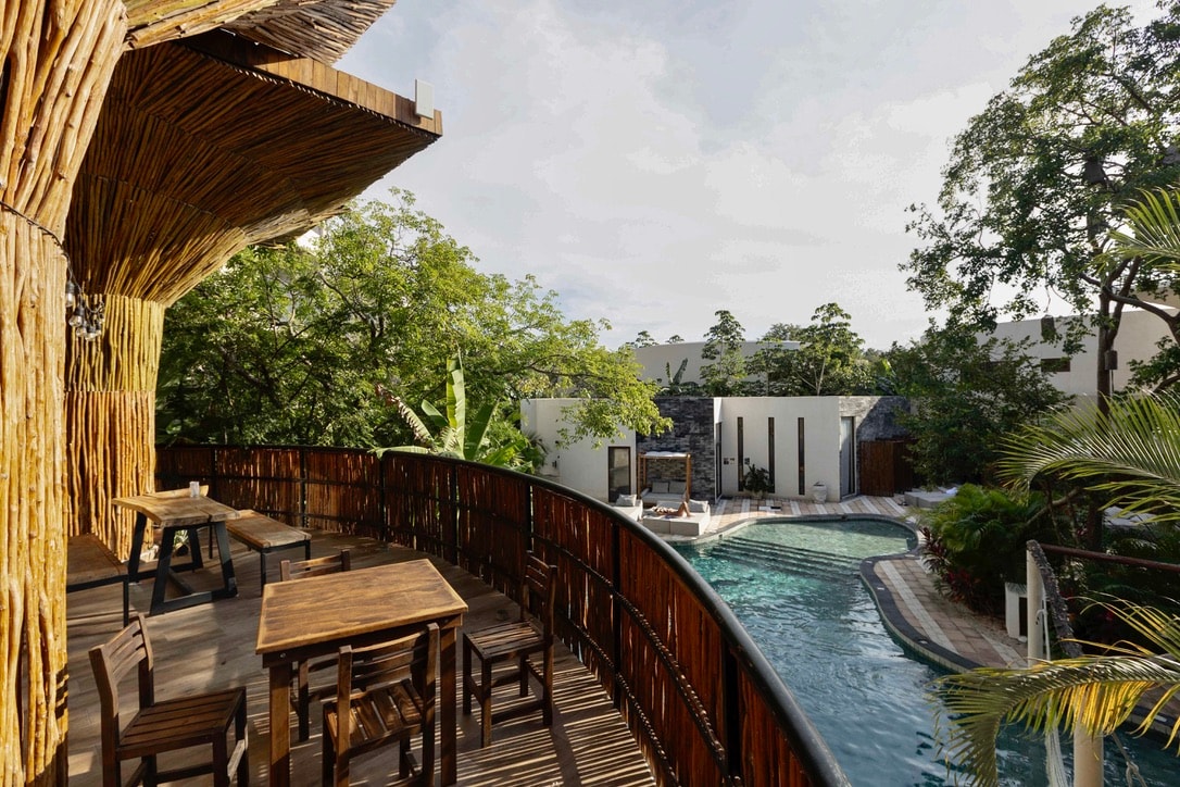 An outdoor deck area features wooden furniture arranged around a railing, overlooking a tranquil pool surrounded by lush greenery. Sunlight filters through the leaves, creating a serene atmosphere. The pool area includes comfortable lounge spots for relaxation, blending seamlessly into the natural surroundings.
