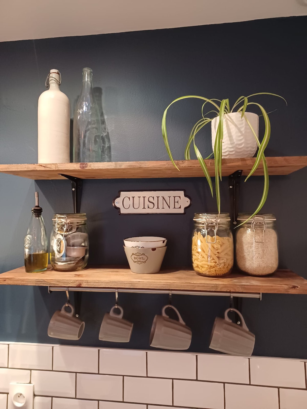 A modern kitchen shelf displays neatly organized jars filled with pasta, rice, and utensils. A decorative sign labeled 'CUISINE' is centered between the items. Glass bottles are positioned to one side, while a small plant adds a refreshing touch. Several hanging mugs are visible below.