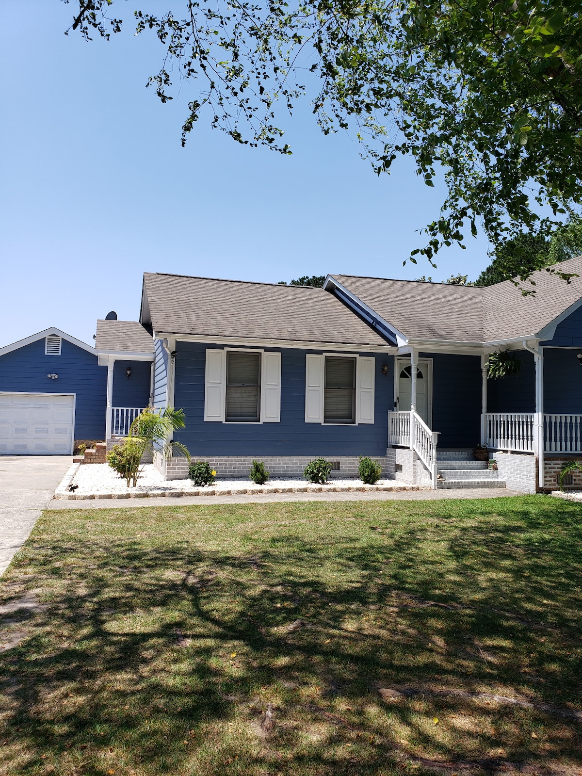 A blue single-story house is featured, adorned with white shutters and a welcoming front porch. A well-maintained lawn is visible, surrounded by subtle landscaping. A garage structure is positioned nearby, complemented by a clear sky.