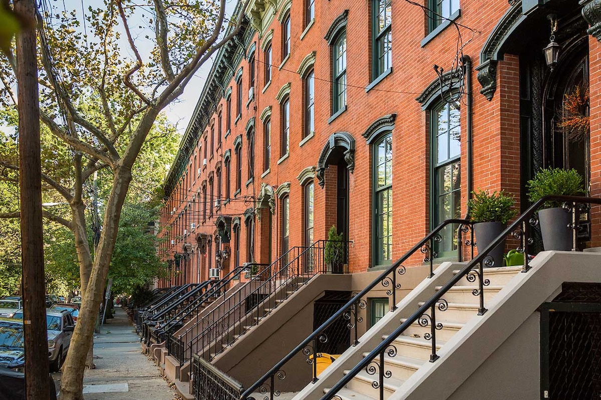 The image showcases a row of classic brick-townhouses featuring ornate architecture. Each home has a distinct staircase leading up to the entrance, lined with greenery and bordered by trees. Sunlight filters through the leaves, creating a peaceful residential ambiance.