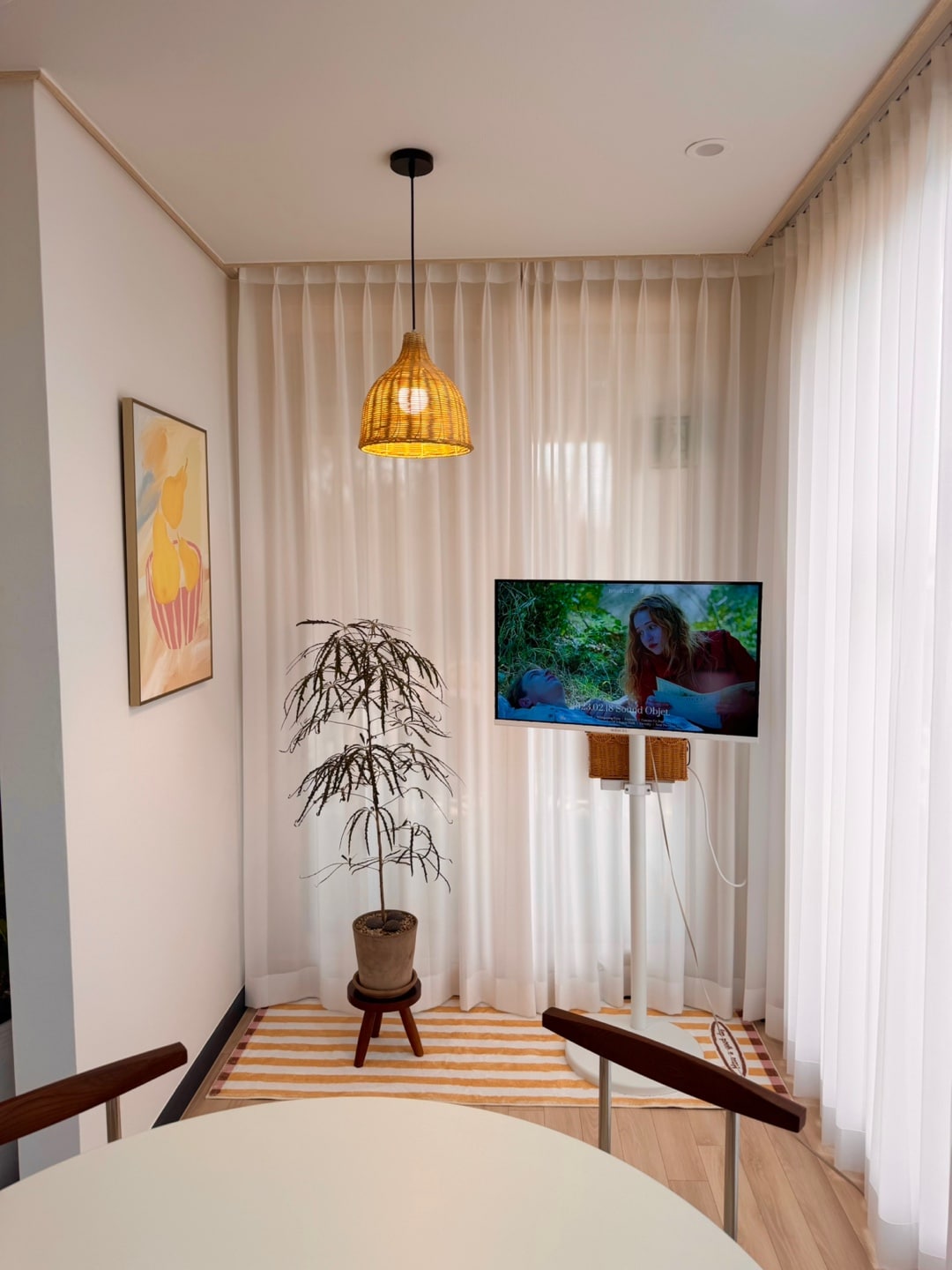 A cozy corner of the living area features a simple table and wooden chairs, accompanied by a potted plant. A flat-screen television is mounted on the wall, displaying a nature scene. Sheer curtains allow soft light to filter in, enhancing the inviting atmosphere.