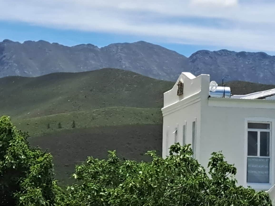 The image captures a corner of a white building framed by lush green foliage, with a backdrop of rolling hills and mountains. The clear blue sky extends above the scene, highlighting the natural beauty surrounding the property.