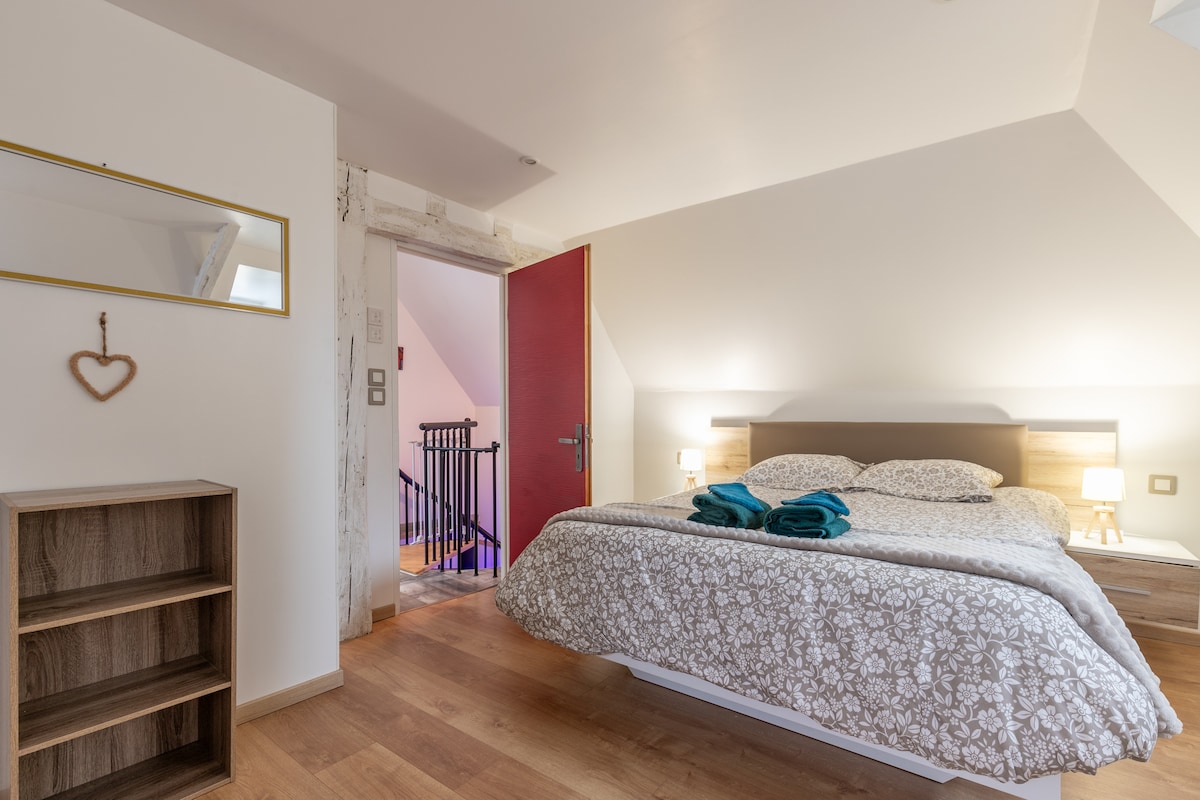 A well-lit bedroom features a spacious bed adorned with a patterned duvet and neatly arranged towels. A full-length mirror is mounted on the wall, reflecting the room's design. Soft bedside lamps provide gentle illumination, while a vibrant red door adds a pop of color.