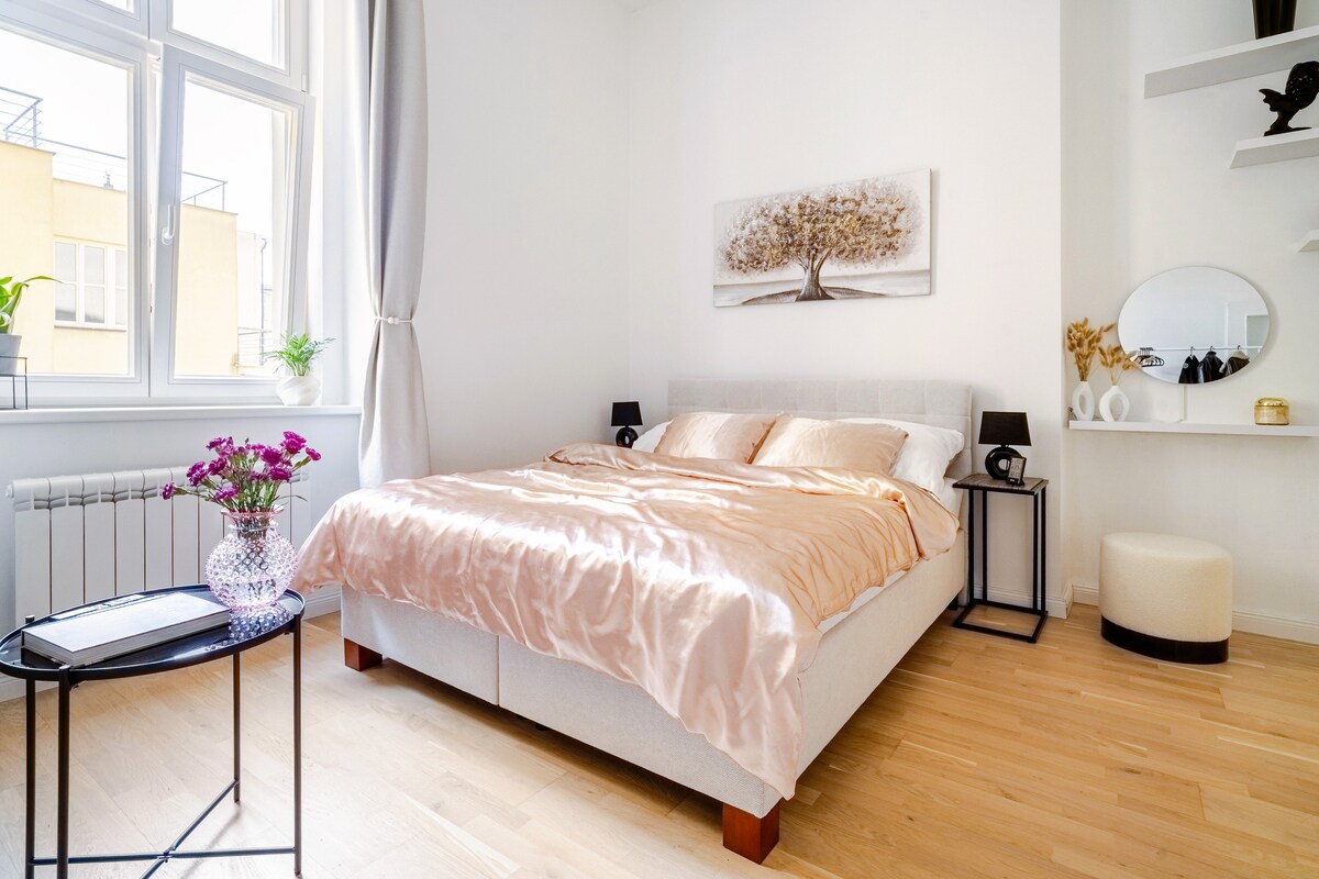 A bright and airy bedroom features a spacious bed dressed in neutral tones. Natural light filters through large windows, illuminating a side table adorned with a vase of flowers. A simple shelf and a round mirror add to the minimalist decor.