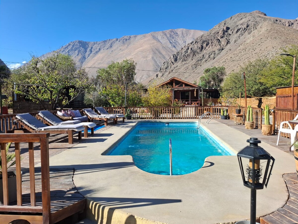 A serene swimming pool is situated in a sunlit courtyard, surrounded by comfortable lounge chairs. Lush greenery and rustic wooden structures are visible in the background, with majestic mountains rising up, contributing to the tranquil ambiance of the space.