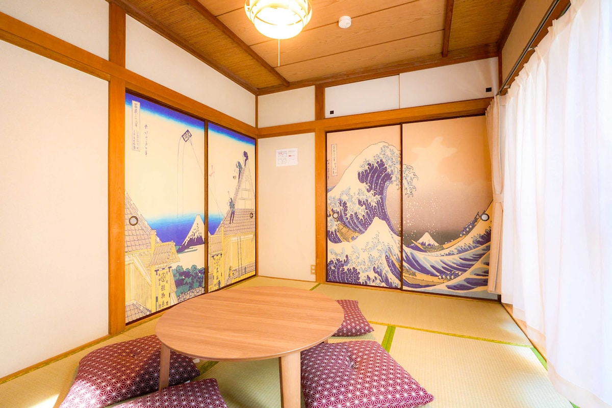 The room features traditional sliding doors adorned with scenic art, including waves and landscapes. A low wooden table is surrounded by tatami mats, with floor cushions arranged for seating. Soft natural light filters through the white curtains, enhancing the serene atmosphere.