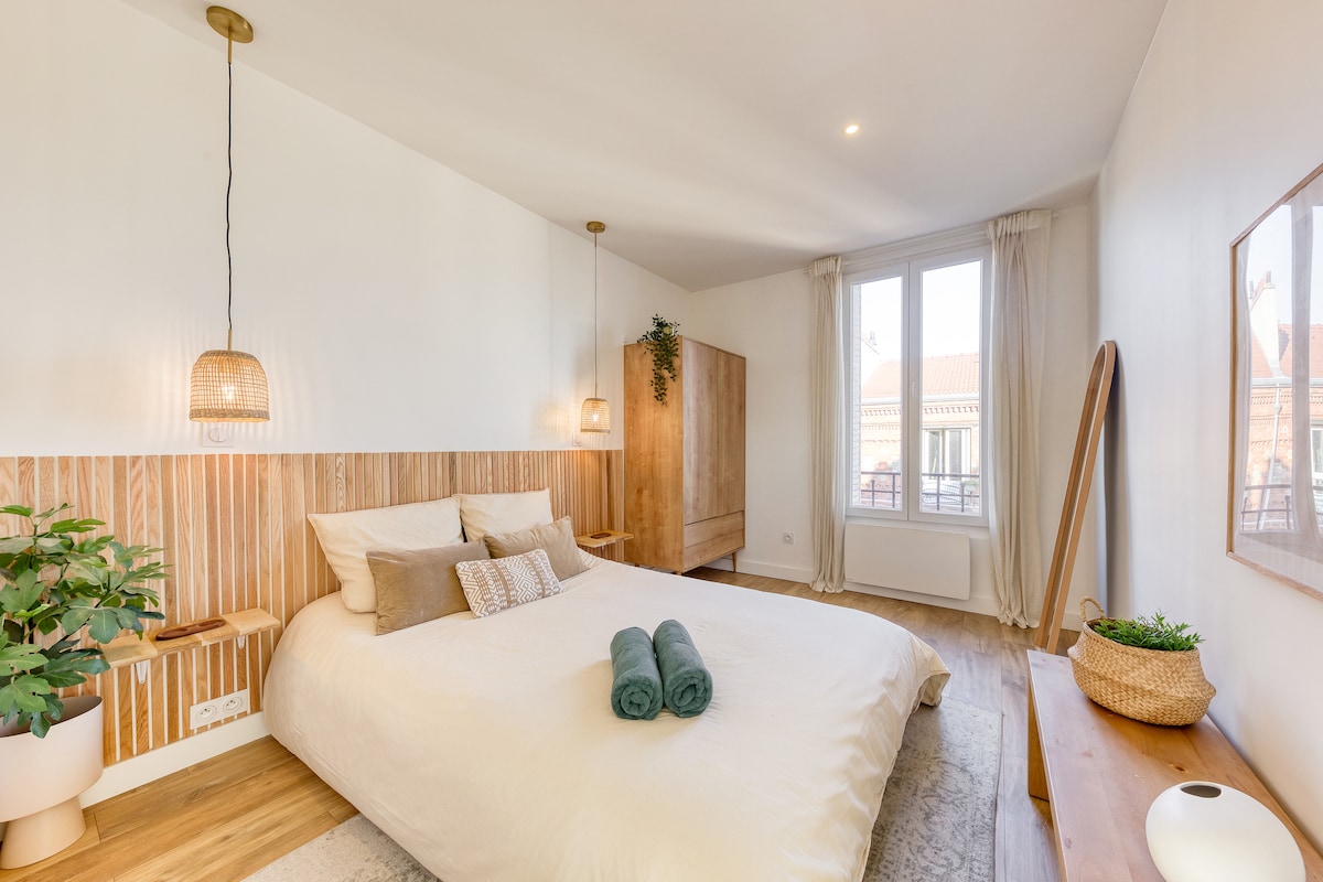 A spacious bedroom features a comfortable double bed adorned with soft bedding and accent pillows. Two pendant lights hang above the bed, while a mirrored cabinet and a decorative plant add warmth to the space. Natural light floods in through large windows, enhancing the serene atmosphere.