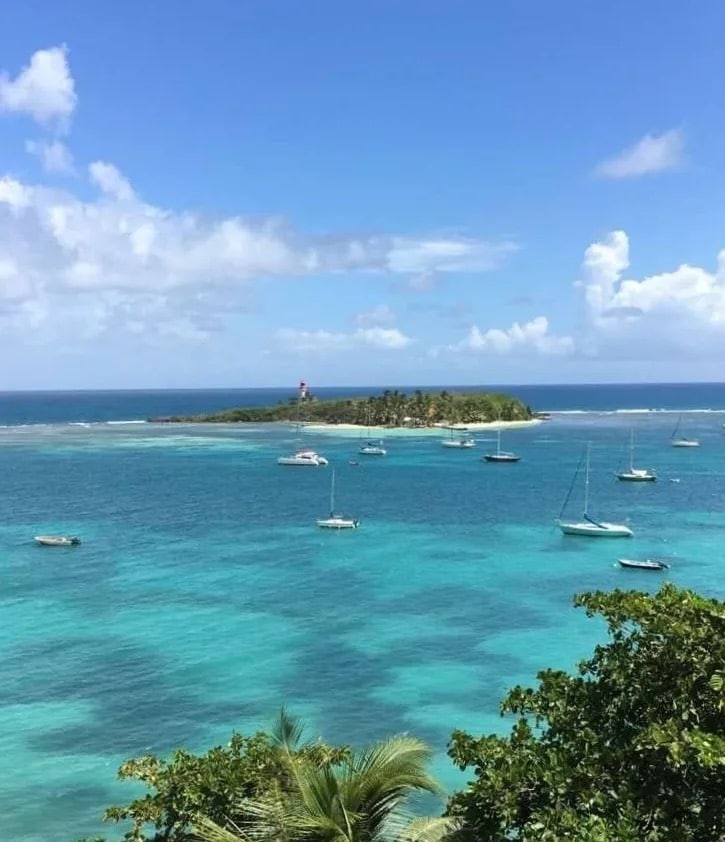 An expansive view of a tranquil turquoise sea, dotted with various sailboats. In the distance, a small island features lush greenery and a lighthouse, under a clear blue sky adorned with soft white clouds.