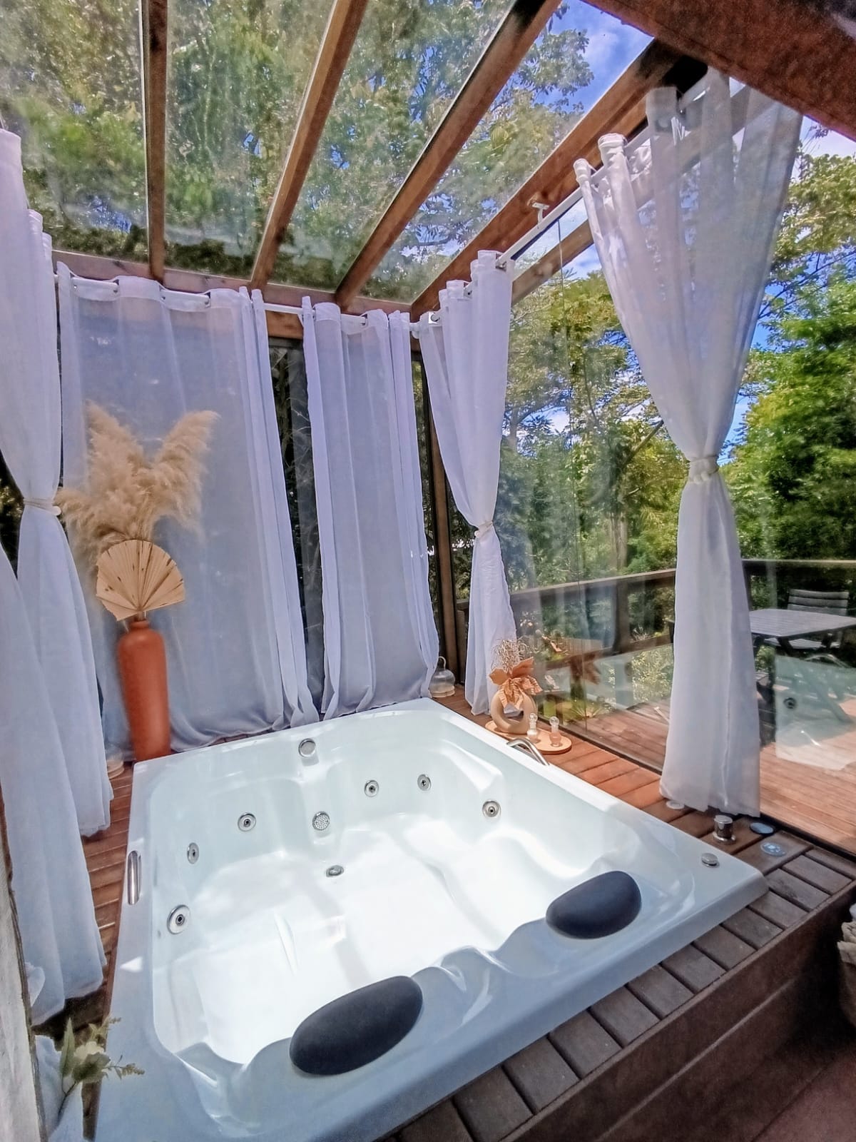 A private indoor hot tub, enclosed by large windows and sheer white curtains, creates a serene ambiance. Natural greenery is visible just outside, enhancing the tranquil setting. Light filters in, reflecting off the water's surface, inviting relaxation in a peaceful atmosphere.
