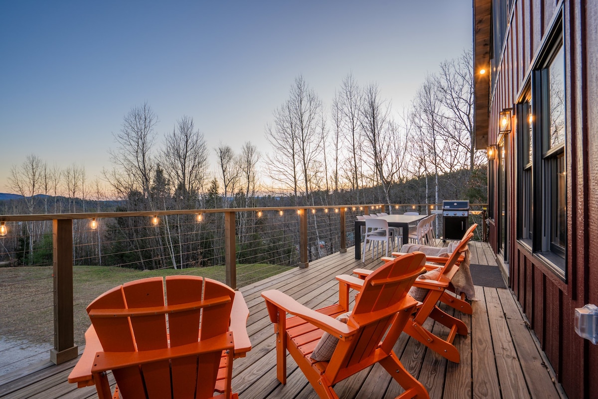 A spacious outdoor deck is furnished with multiple orange Adirondack chairs, inviting relaxation. Twinkling string lights add ambiance, while a scenic view of trees and mountains creates a serene backdrop. The deck accommodates a BBQ grill and a dining area, enhancing the outdoor experience.
