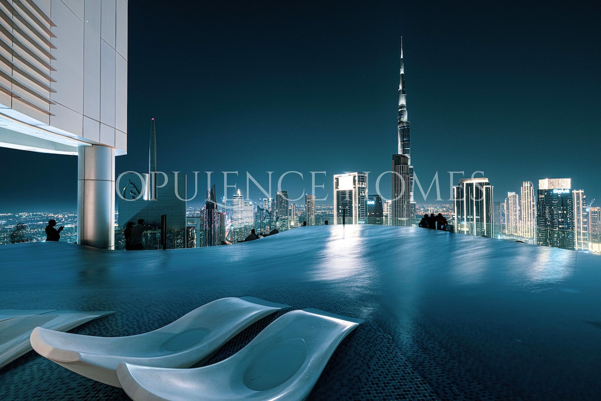 An infinity pool on the rooftop offers stunning views of the Dubai skyline at night, with the Burj Khalifa prominently visible. Relaxation loungers are positioned at the edge of the pool, allowing for an uninterrupted view of the shimmering city lights.