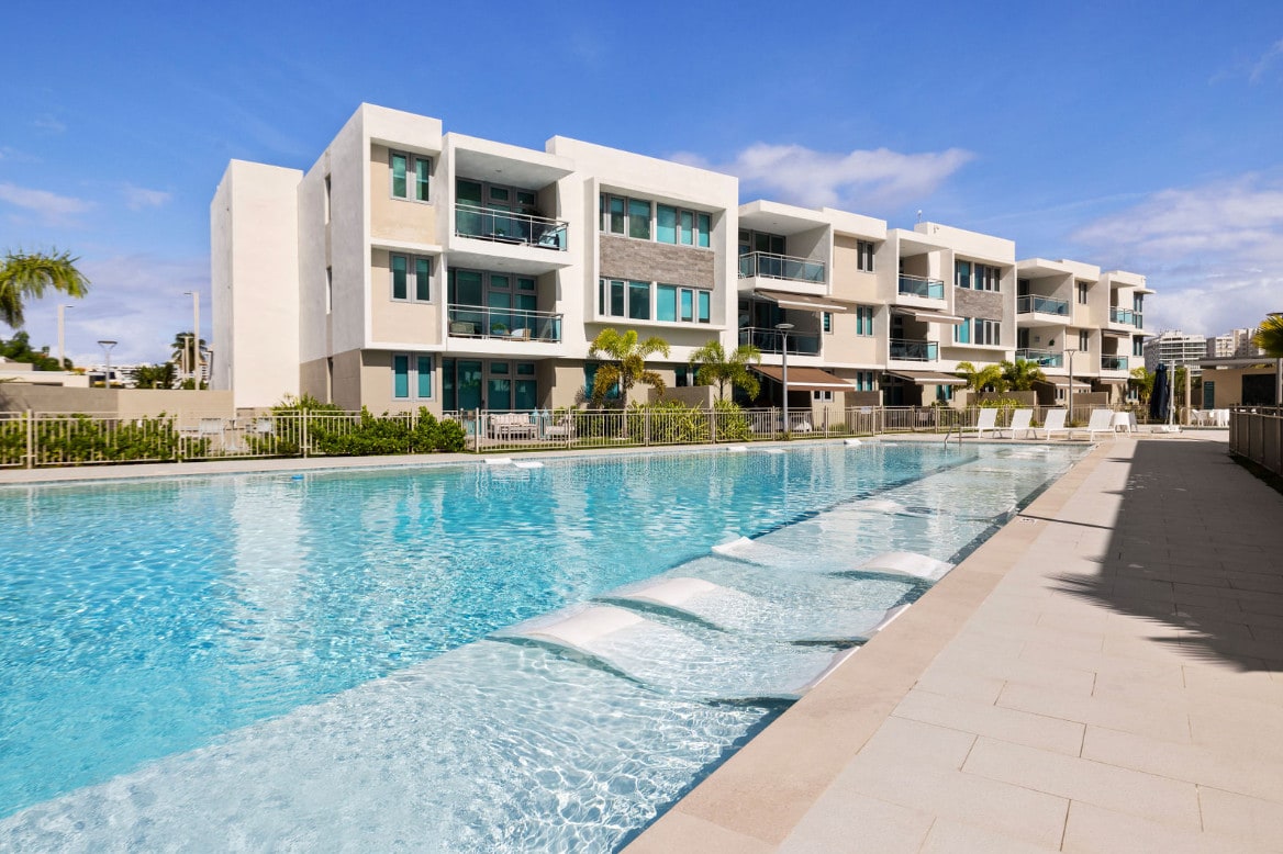A serene swimming pool stretches alongside modern residences. The clear blue water reflects the sky, with gentle water features providing a calming effect. Lush greenery frames the area, creating a clean and inviting space for relaxation and recreation.