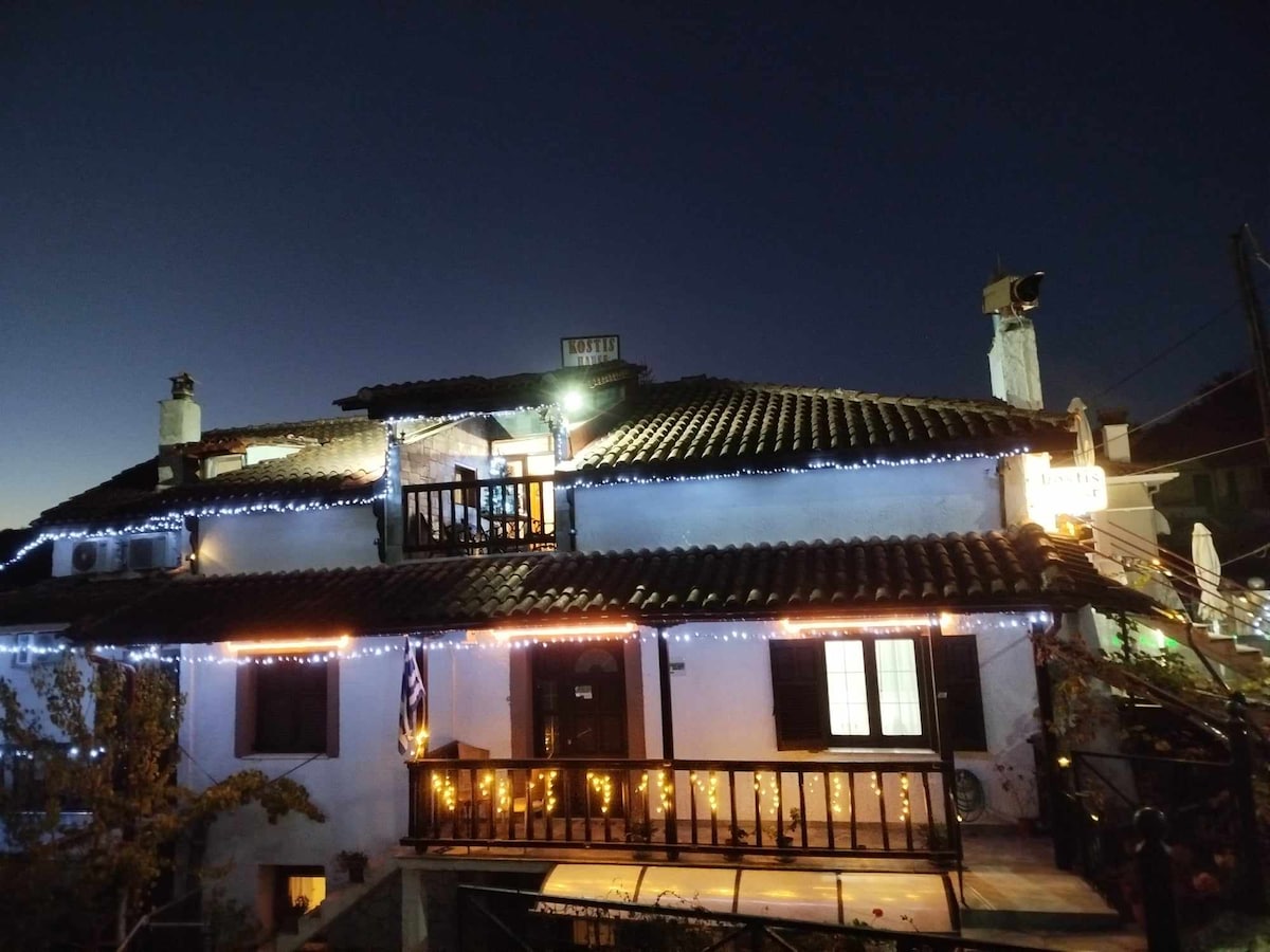 A two-story building with a rustic facade is illuminated by twinkling lights at dusk. Outdoor balconies exhibit wooden railings, while multiple windows reflect a warm glow from within. The structure is surrounded by greenery, adding to the charming ambiance of the setting.