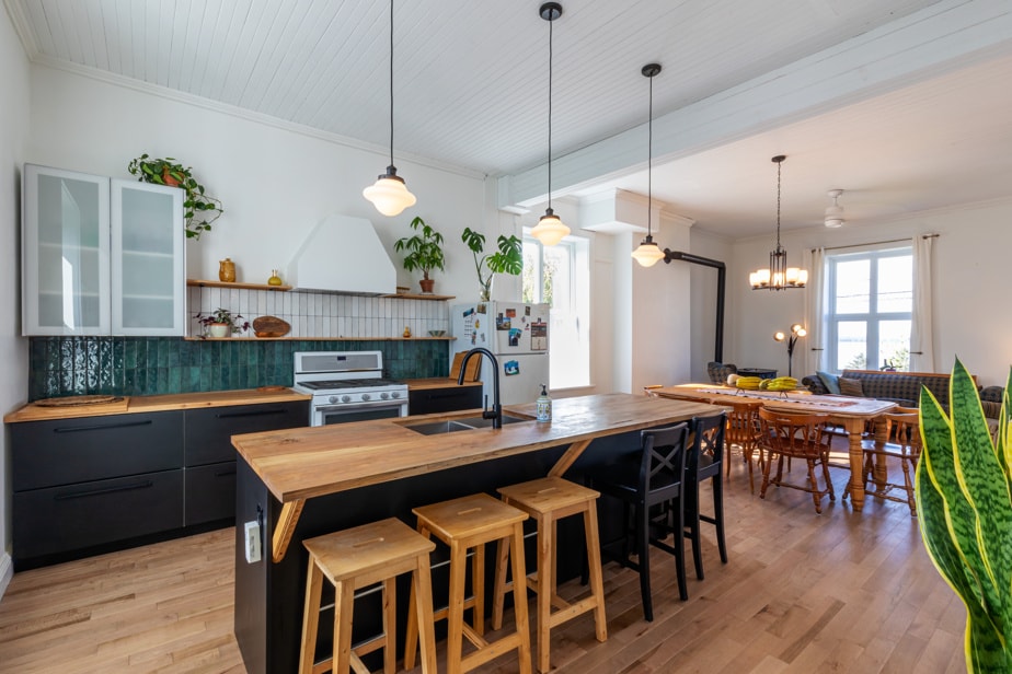 A spacious kitchen features wooden countertops and a central island with seating. Sleek cabinetry and a modern stove complement the design. Natural light fills the room from multiple windows, enhancing the inviting atmosphere. A dining area with wooden chairs is visible in the background.