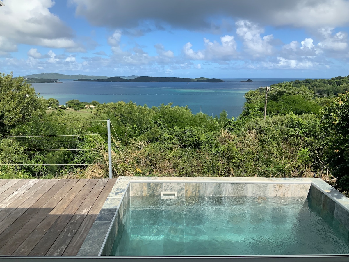 A view of a small pool reveals tranquil waters overlooking lush greenery and the expansive turquoise sea. The horizon features gentle waves and distant islets, with a clear sky dotted with soft clouds. The wooden deck enhances the connection to the serene outdoor environment.
