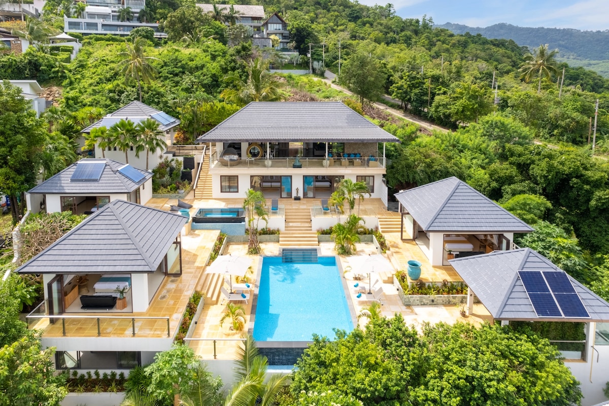An aerial view captures the villa surrounded by lush greenery, featuring multiple structures with contemporary architecture. A large swimming pool is centrally located, framed by palm trees and spacious terraces. Each building is connected by outdoor stairs, enhancing accessibility and flow.