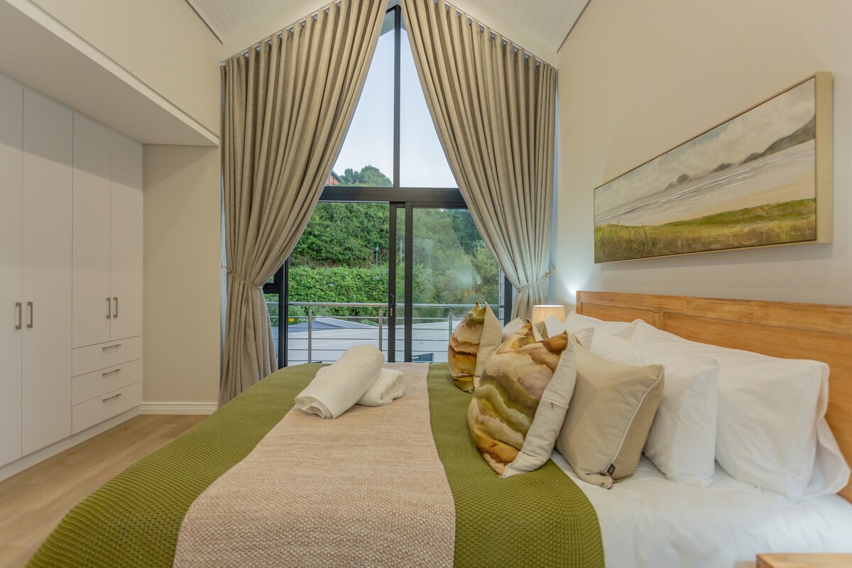 The elegant bedroom features a spacious bed adorned with soft, neutral-toned pillows and a textured throw. Large windows with flowing curtains invite natural light and offer views of the surrounding greenery. Built-in wardrobes complement the modern design, providing ample storage.