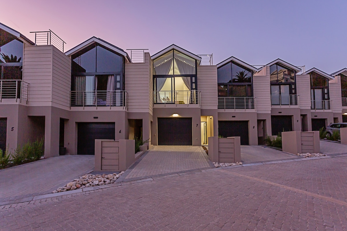 A modern two-story building is presented, showcasing a row of stylish apartments with large windows and a distinctive triangular roofline. Bright outdoor lighting and landscaped pathways create an inviting atmosphere. The neatly arranged stones along the pavement add a touch of elegance to the exterior.