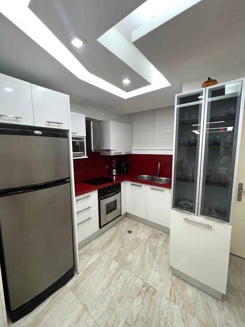 A modern kitchen is highlighted by sleek white cabinetry and a contrasting red backsplash. Stainless steel appliances include a refrigerator and built-in oven. The space is illuminated by recessed lighting, and a glass cabinet showcases dishware. Marble flooring contributes to a polished appearance.