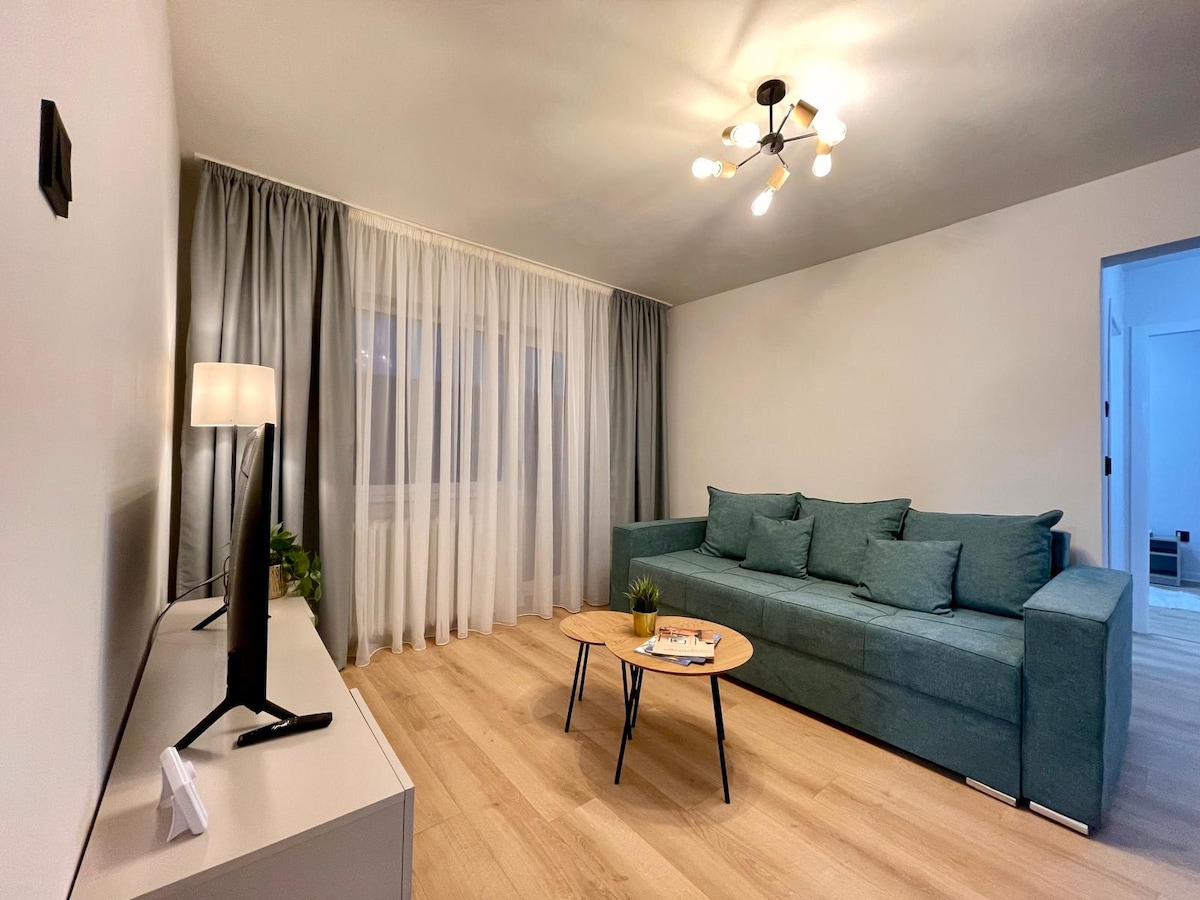 A comfortable living area showcases a green sofa and a light wood coffee table. Natural light filters through sheer curtains, highlighting the modern design. A television is positioned on a sleek console, complemented by a small plant adding a touch of greenery to the space.