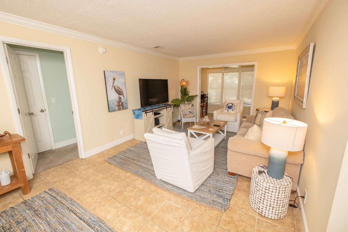 A light-filled living area features a comfortable beige sofa and two white chairs positioned around a coffee table. A flat-screen TV is mounted on the wall, and a lamp provides soft illumination. A door leads to an outdoor area, enhancing the spacious ambiance.