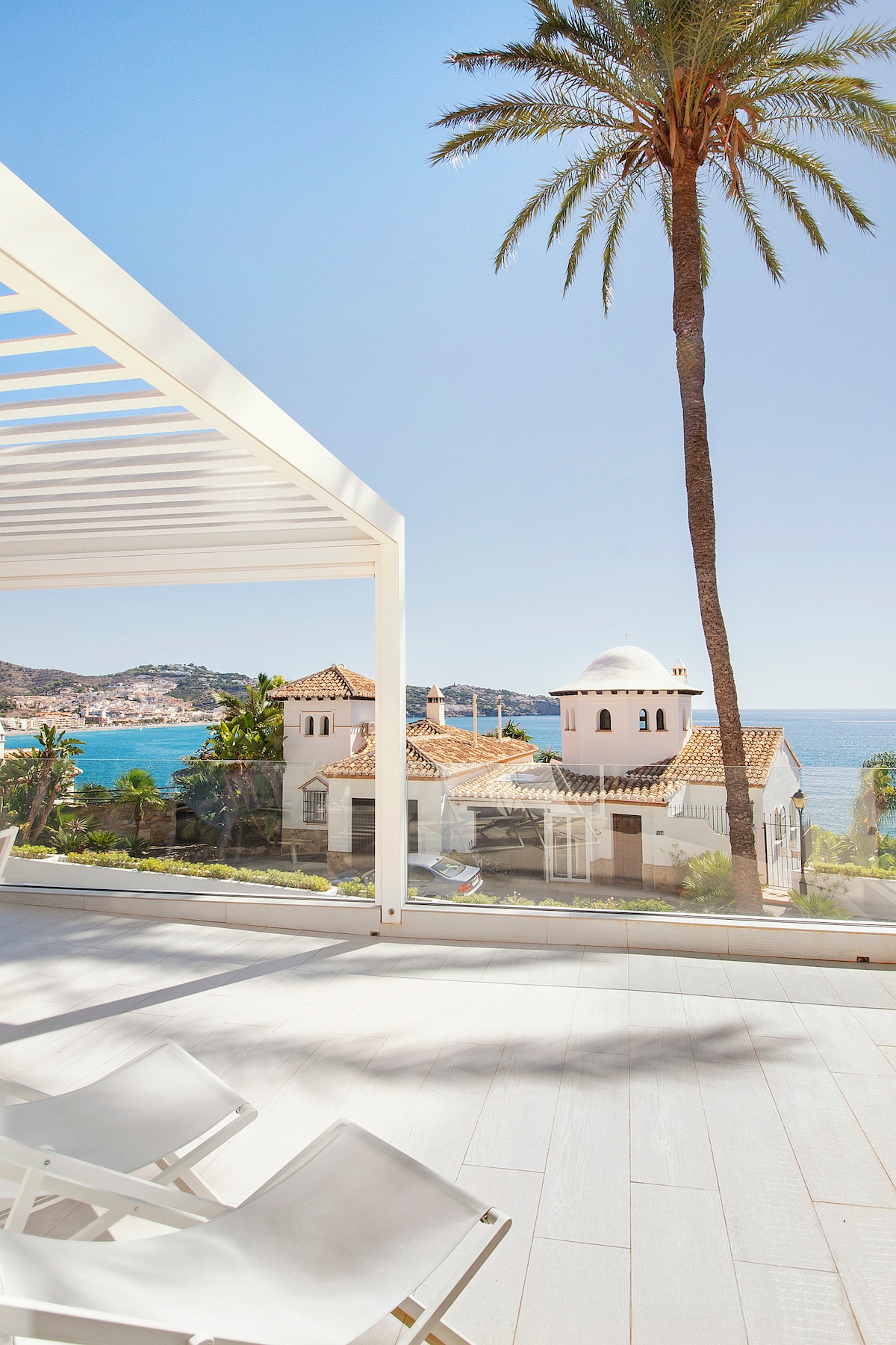 A spacious outdoor terrace features a modern design with a clear view of the sea and nearby palm trees. Lounge chairs are positioned to enjoy the scenic coastal landscape, which includes charming architecture and lush greenery in the background.