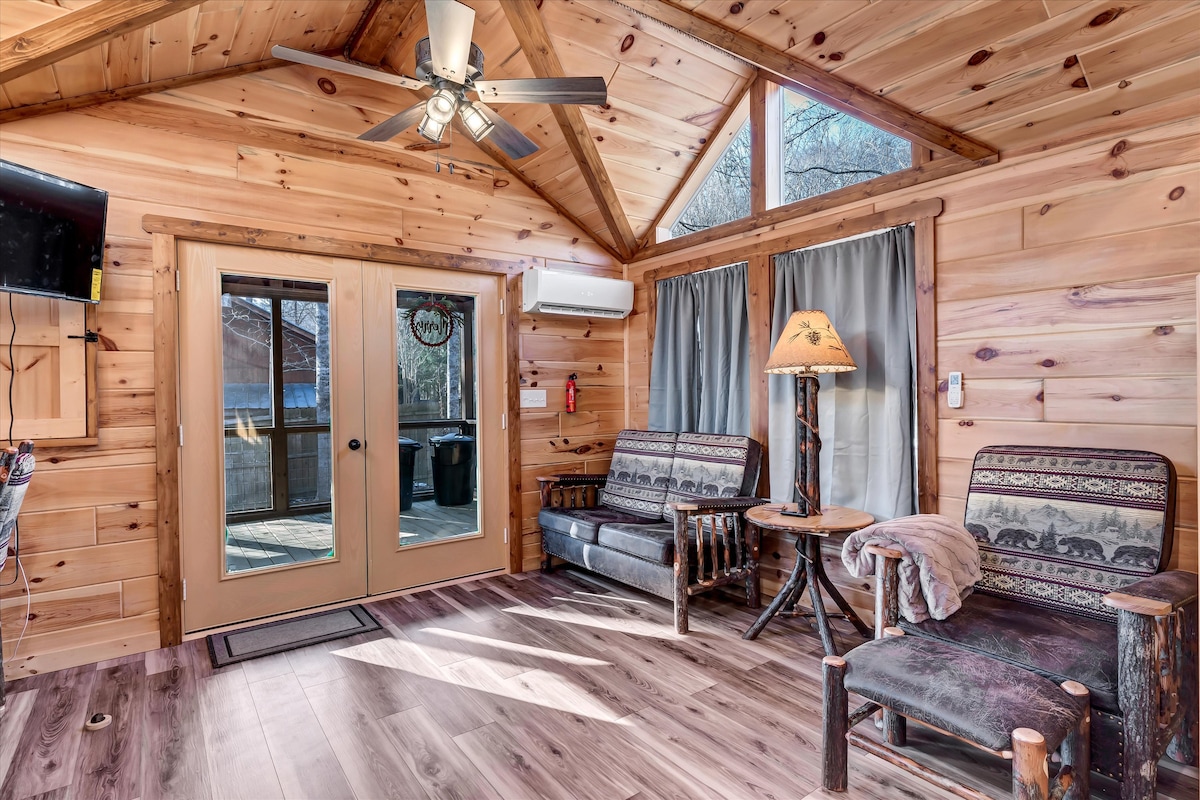 A cozy living area features wood-paneled walls and vaulted ceilings with a ceiling fan. Two rustic sofas and a wooden chair surround a small table, while a door leads to an outdoor space. Natural light fills the room through large windows, accented with soft curtains.